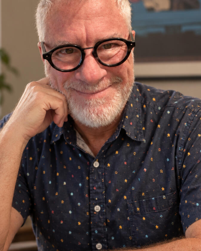 Arden McKenna smiling at a writing desk, wearing round black glasses and a navy patterned shirt.